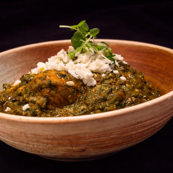 A rustic bowl filled with green curry, topped with crumbled white cheese and fresh herbs.