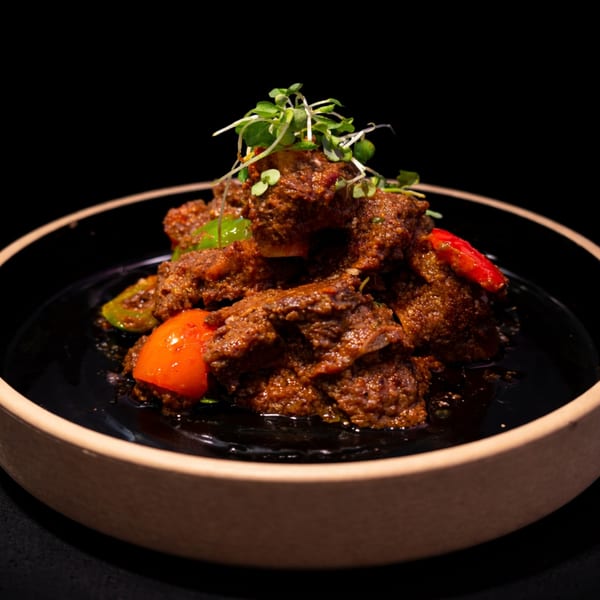 Spicy beef rendang with red and green peppers, garnished with microgreens in a black bowl.