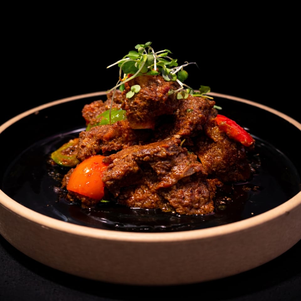 Spicy beef rendang with red and green peppers, garnished with microgreens in a black bowl.
