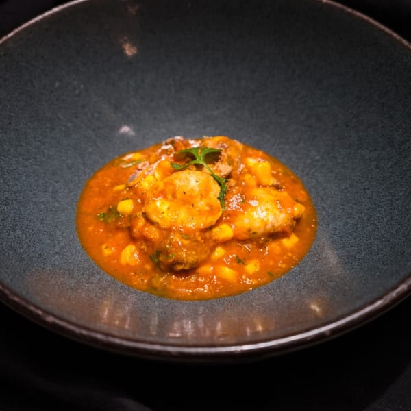 A dark bowl holds a vibrant orange stew with shrimp, corn, and herbs, on a dark surface.